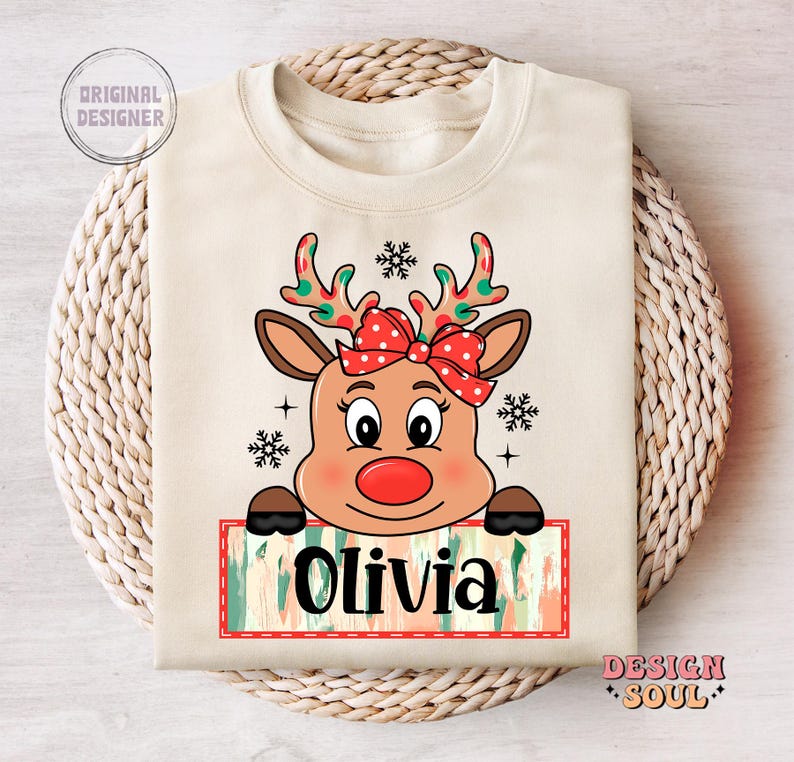 May include: A beige sweatshirt with a cartoon reindeer wearing a red bow. The reindeer is looking at the viewer and has a red nose. The text "Olivia" is written on a colorful banner below the reindeer.
