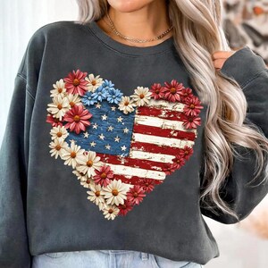 May include: Dark gray sweatshirt featuring a heart-shaped design of the American flag with a distressed wood effect. The flag is surrounded by red, white, and blue flowers, creating a patriotic and floral theme.
