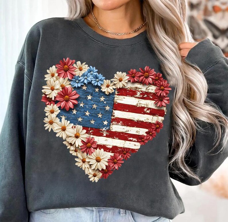 May include: Dark gray sweatshirt featuring a heart-shaped design of the American flag with a distressed wood effect. The flag is surrounded by red, white, and blue flowers, creating a patriotic and floral theme.