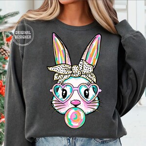 May include: A dark grey sweatshirt featuring a colourful graphic of a bunny wearing pink heart-shaped sunglasses and a polka dot headband. The bunny is blowing a pink and blue bubble.