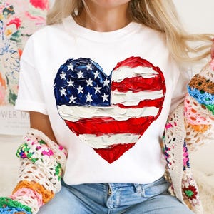 Pu&ograve; includere: Maglietta bianca con un design a forma di cuore dipinto a mano con i colori della bandiera americana. La bandiera &egrave; rossa, bianca e blu.