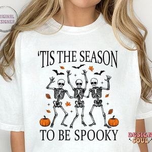 May include: White t-shirt with a black graphic design featuring three skeletons dancing with the text 'Tis the season to be spooky' and two pumpkins.