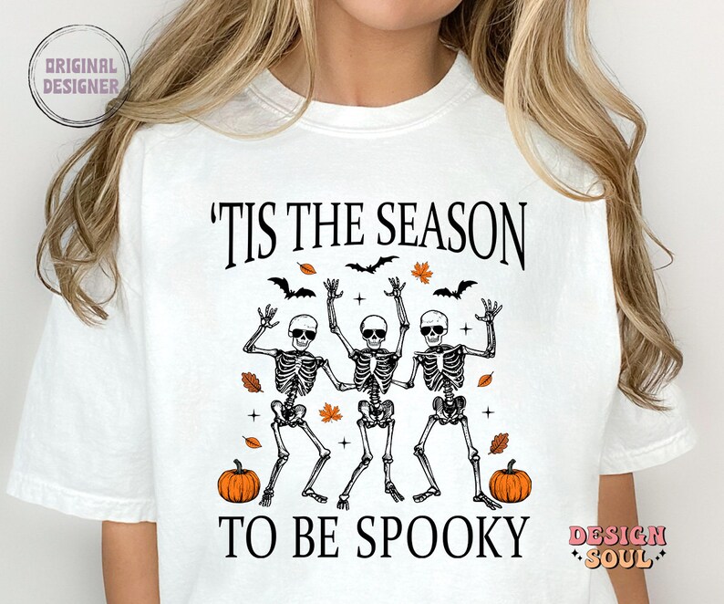 May include: White t-shirt with a black graphic design featuring three skeletons dancing with the text 'Tis the season to be spooky' and two pumpkins.