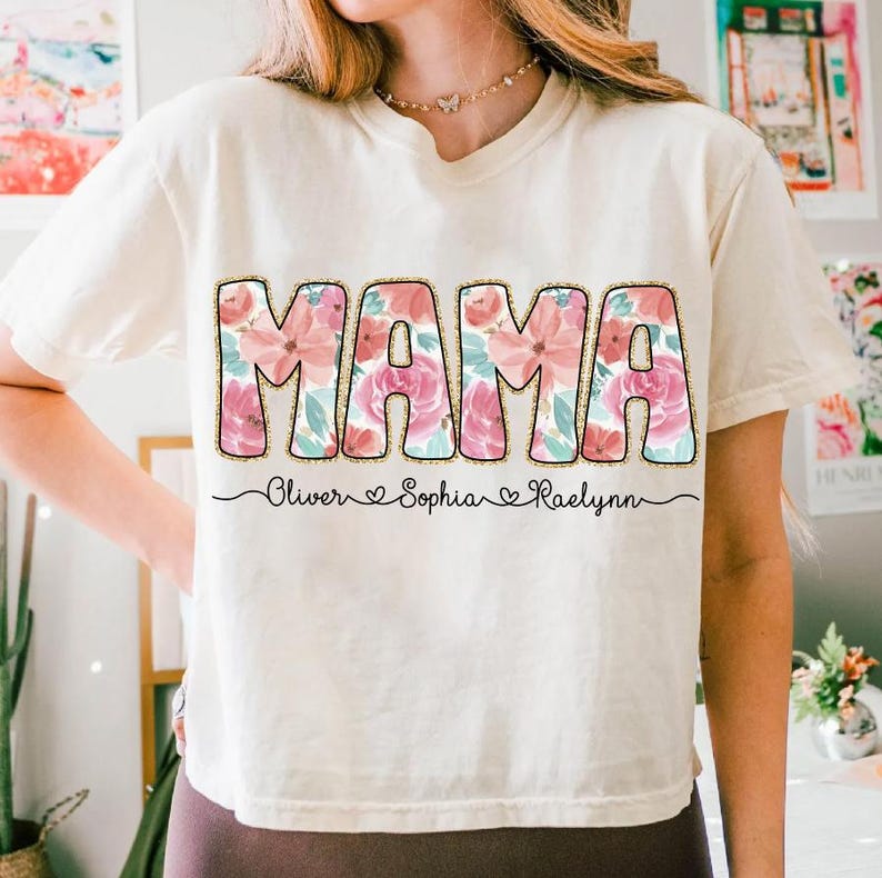 May include: A white t-shirt with the word "MAMA" in large, pink floral letters. The letters are outlined in gold glitter. Below the word "MAMA" is the text "Oliver Sophia Raelynn" with hearts between the names.