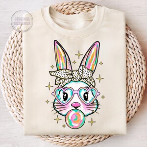 May include: A white sweatshirt with a colorful cartoon bunny wearing pink heart-shaped sunglasses and a black and white polka dot headband. The bunny is holding a colorful lollipop in its mouth.