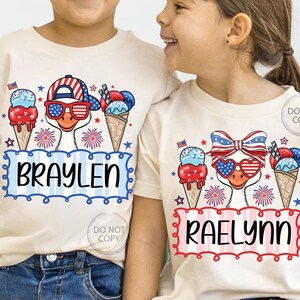 May include: Two children's t-shirts with patriotic ice cream cone designs. One shirt features the name "BRAYLEN" in blue, the other "RAELYNN" in red. Each shirt has a cartoon duck wearing sunglasses and a hat with a red, white, and blue theme.