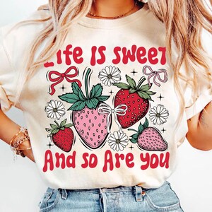 May include: A white t-shirt with a graphic design of strawberries and flowers. The text "Life is sweet and so are you" is printed in red on the shirt.