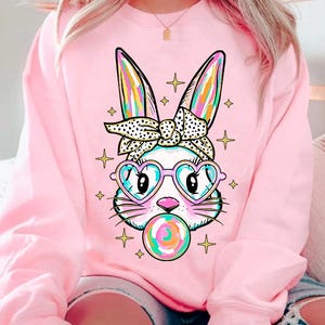 May include: Pink sweatshirt with a graphic of a white bunny wearing heart-shaped sunglasses and a polka dot headband, blowing a bubblegum bubble. The bunny has rainbow-colored ears and the bubblegum is multi-colored.