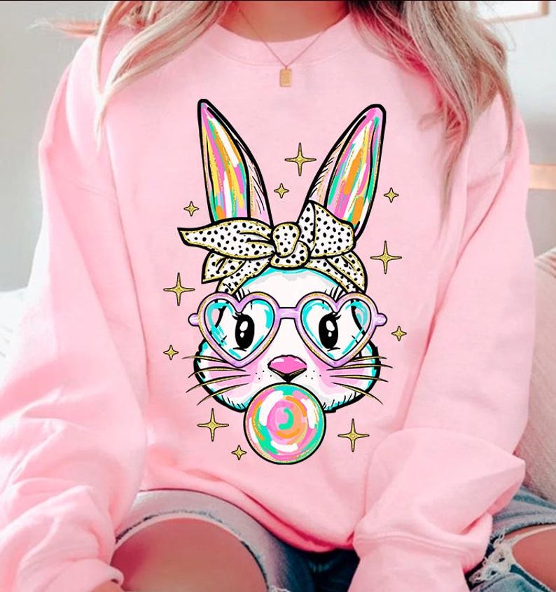 May include: Pink sweatshirt with a graphic of a white bunny wearing heart-shaped sunglasses and a polka dot headband, blowing a bubblegum bubble. The bunny has rainbow-colored ears and the bubblegum is multi-colored.