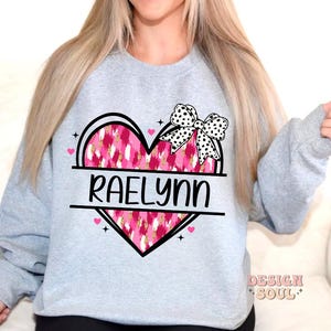 May include: A light grey sweatshirt with a pink and white heart design. The heart has a black and white bow on top and the name "Raelynn" written in black letters.