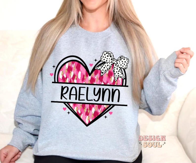 May include: A light grey sweatshirt with a pink and white heart design. The heart has a black and white bow on top and the name "Raelynn" written in black letters.