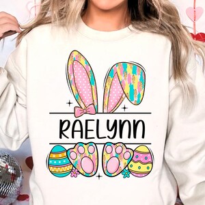 May include: A white sweatshirt with a colorful Easter bunny design. The bunny ears are pink, yellow, and blue with white polka dots. The bunny has a pink bow and paw prints. The text "Raelynn" is printed below the bunny. The sweatshirt also features three colorful Easter eggs.
