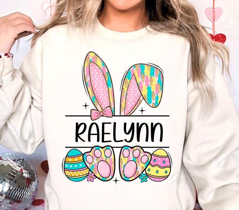 May include: A white sweatshirt with a colorful Easter bunny design. The bunny ears are pink, yellow, and blue with white polka dots. The bunny has a pink bow and paw prints. The text "Raelynn" is printed below the bunny. The sweatshirt also features three colorful Easter eggs.