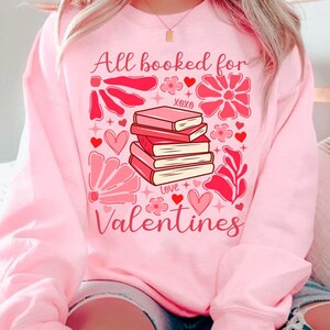 May include: Pink sweatshirt with a graphic design of a stack of books, hearts, flowers, and the text "All booked for Valentines".