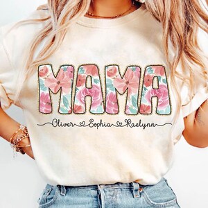 May include: A white t-shirt with a floral design that says "MAMA" in pink and gold letters. The shirt also has the names "Oliver", "Sophia", and "Raelynn" written below the word "MAMA".