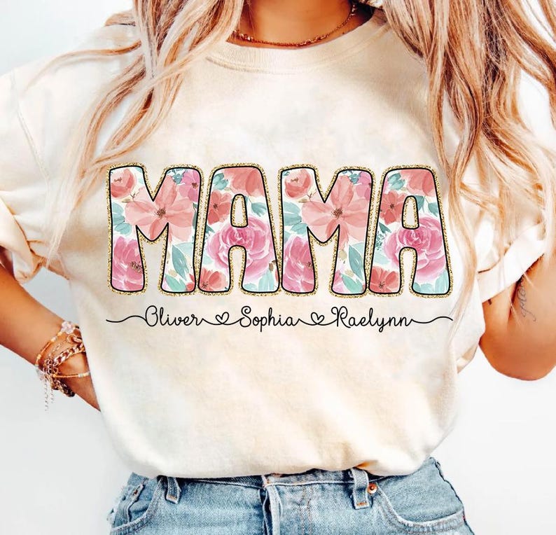 May include: A white t-shirt with a floral design that says "MAMA" in pink and gold letters. The shirt also has the names "Oliver", "Sophia", and "Raelynn" written below the word "MAMA".