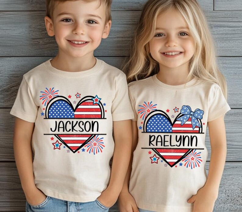 May include: Two children's t-shirts with a heart-shaped design featuring the American flag. One shirt has the name "JACKSON" and the other has "RAELYNN". The design includes fireworks and stars in red, white, and blue.