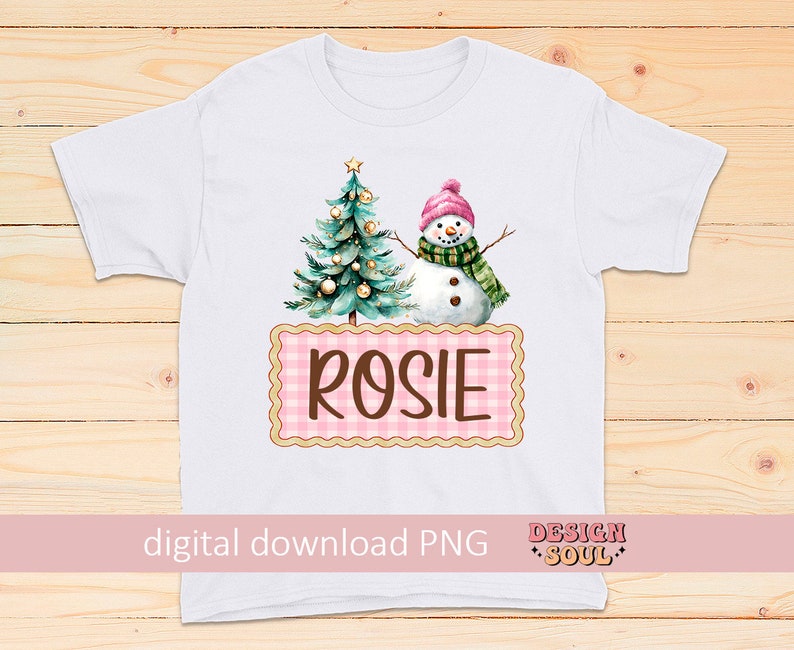 May include: White t-shirt with a pink and white gingham design featuring a snowman and a Christmas tree with the name "Rosie" on it.