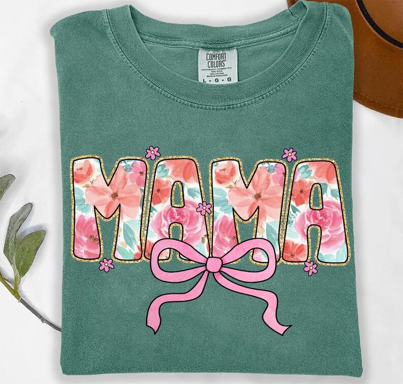 May include: A green t-shirt with a floral design that says "MAMA" in gold glitter letters with a pink bow underneath.