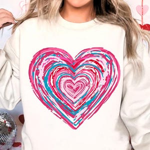 Pu&ograve; includere: Una felpa bianca con un grande disegno a forma di cuore rosa, rosso e blu. Il cuore &egrave; composto da cerchi concentrici di colore.