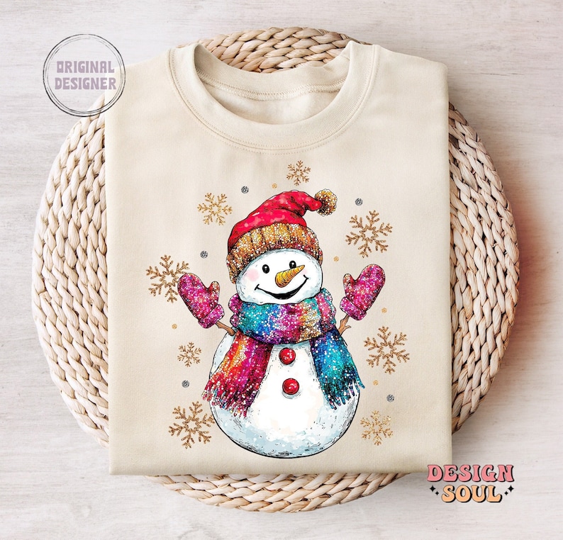May include: A beige sweatshirt with a cartoon snowman wearing a red and white hat, a colorful scarf, and pink mittens. The snowman is surrounded by gold glitter snowflakes.