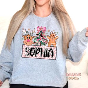 Op de afbeelding: Een grijs sweatshirt met een roze en groen kerstontwerp met een rendier, een peperkoekmannetje en een kerstboom. Het ontwerp bevat ook de naam "Sophia" in zwarte tekst.