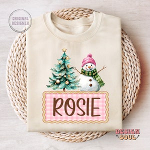 May include: A cream colored sweatshirt with a watercolor illustration of a Christmas tree and snowman. The text "ROSIE" is written in brown letters on a pink and white gingham background.