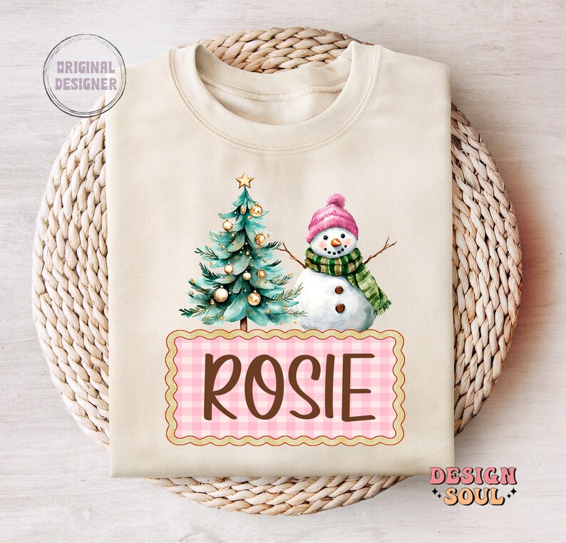 May include: A cream colored sweatshirt with a watercolor illustration of a Christmas tree and snowman. The text "ROSIE" is written in brown letters on a pink and white gingham background.