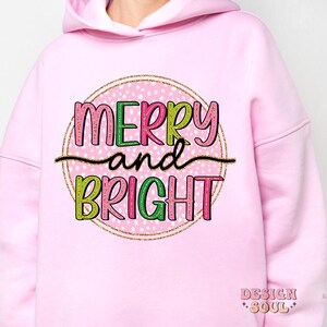 May include: A light pink hoodie with the words "MERRY and BRIGHT" in a colorful, playful font. The text is set within a pink circle with white polka dots and a gold glitter border. The hoodie has a drawstring hood.
