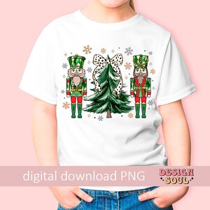 May include: White t-shirt with a festive Christmas design. The design features two nutcrackers in red, green, and gold, a green Christmas tree with a polka dot bow, and snowflakes. The words "digital download PNG" are printed at the bottom.