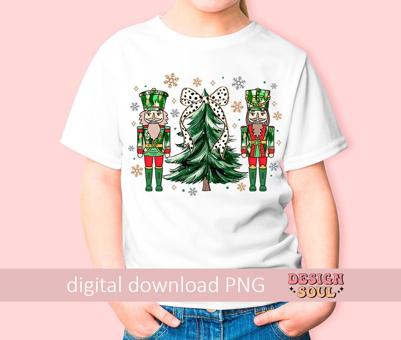 May include: White t-shirt with a festive Christmas design. The design features two nutcrackers in red, green, and gold, a green Christmas tree with a polka dot bow, and snowflakes. The words "digital download PNG" are printed at the bottom.