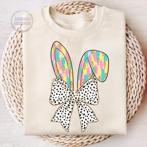 May include: A cream-colored sweatshirt with a graphic of a black and white polka dot bow with pastel pink, yellow, and blue bunny ears.