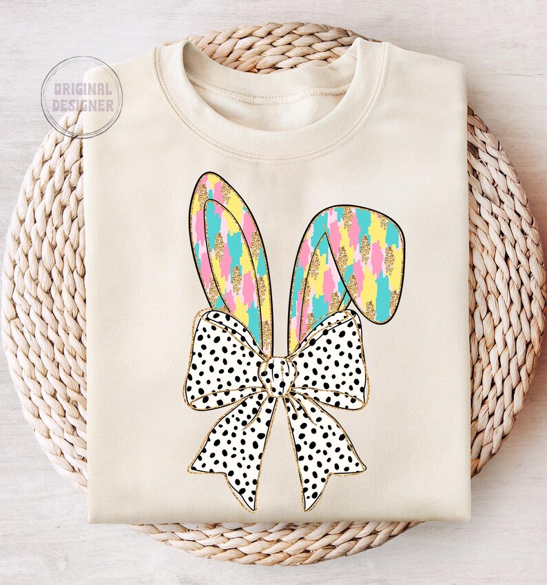 May include: A cream-colored sweatshirt with a graphic of a black and white polka dot bow with pastel pink, yellow, and blue bunny ears.