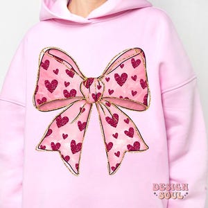May include: Pink hoodie with a large, gold glitter bow with pink hearts. The bow is centered on the front of the hoodie.
