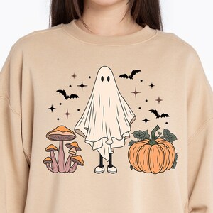May include: A tan sweatshirt with a Halloween-themed design featuring a ghost, mushrooms, and a pumpkin. The ghost is white and has two eyes. The mushrooms are brown with orange tops. The pumpkin is orange with a green stem and leaves.