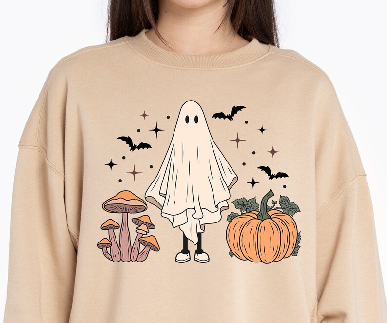 May include: A tan sweatshirt with a Halloween-themed design featuring a ghost, mushrooms, and a pumpkin. The ghost is white and has two eyes. The mushrooms are brown with orange tops. The pumpkin is orange with a green stem and leaves.