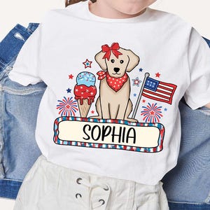 Pu&ograve; includere: Maglietta bianca con un design patriottico rosso, bianco e blu con un cane, un cono gelato e fuochi d'artificio. Il testo "Sophia" &egrave; stampato sulla maglietta.