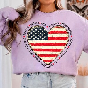 May include: A lavender sweatshirt with a distressed American flag heart design. The heart is surrounded by the words "Home of the Brave Land of the free America Victory USA Freedom Independence Liberty Patriotism Unity".
