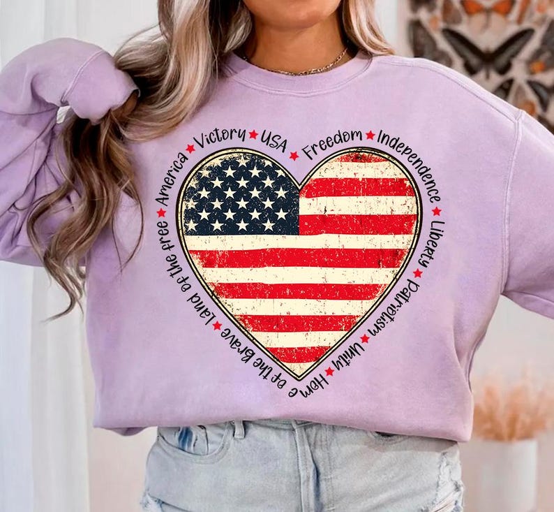 May include: A lavender sweatshirt with a distressed American flag heart design. The heart is surrounded by the words "Home of the Brave Land of the free America Victory USA Freedom Independence Liberty Patriotism Unity".