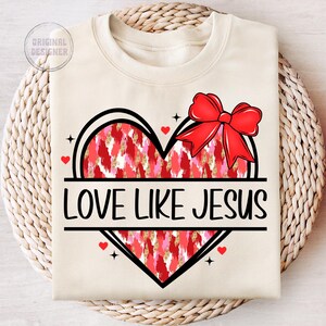 May include: A beige sweatshirt with a red and pink heart design and the text "LOVE LIKE JESUS". The heart has a red bow on top.