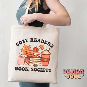 May include: A white tote bag with the text "Cozy Readers Book Society" in brown lettering. The design features a stack of books, a pumpkin, a candle, and a cup of coffee with whipped cream. The text "Design Soul" is printed in pink and white on the bottom right corner of the bag.