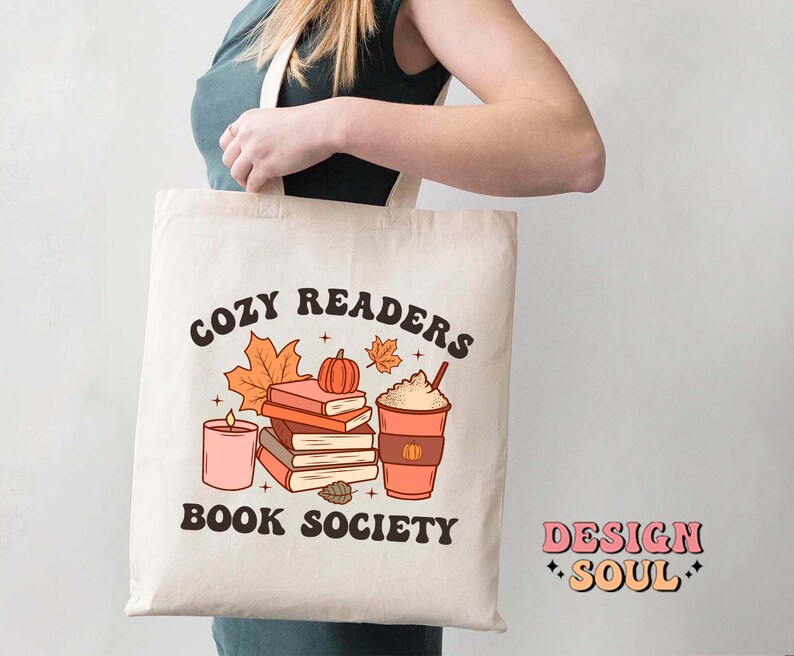 May include: A white tote bag with the text "Cozy Readers Book Society" in brown lettering. The design features a stack of books, a pumpkin, a candle, and a cup of coffee with whipped cream. The text "Design Soul" is printed in pink and white on the bottom right corner of the bag.