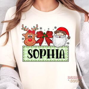 May include: A white long-sleeved shirt with a green and white checkered banner that says "SOPHIA". The banner has a cartoon reindeer with a red bow, a red bow with green holly, and a cartoon Santa Claus with a white beard.