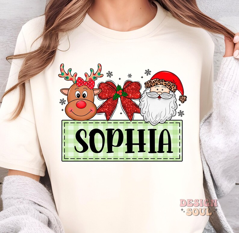 May include: A white long-sleeved shirt with a green and white checkered banner that says "SOPHIA". The banner has a cartoon reindeer with a red bow, a red bow with green holly, and a cartoon Santa Claus with a white beard.