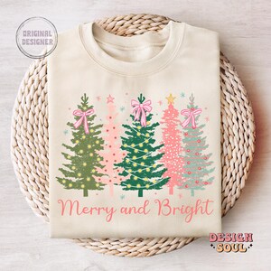 May include: A cream-colored sweatshirt with a festive design of four Christmas trees with pink bows and lights. The text "Merry and Bright" is printed below the trees.