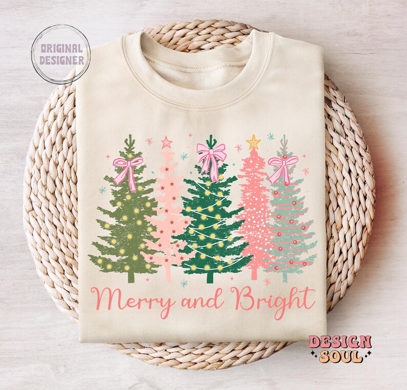 May include: A cream-colored sweatshirt with a festive design of four Christmas trees with pink bows and lights. The text "Merry and Bright" is printed below the trees.