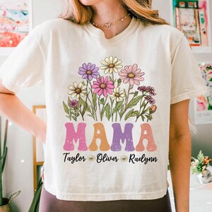 May include: Cream-colored t-shirt featuring a floral design with the word "MAMA" in retro font. Below the word "MAMA" are the names "Taylor", "Oliver", and "Raelynn" with flower icons. The floral design includes daisies in shades of purple, pink, and yellow.