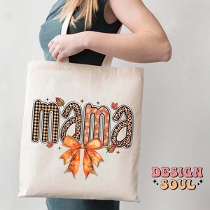 May include: A white tote bag with the word "mama" printed in brown, black, and pink patterned letters. The letters are surrounded by small orange, brown, and black leaves. A large orange bow is centered below the word "mama".