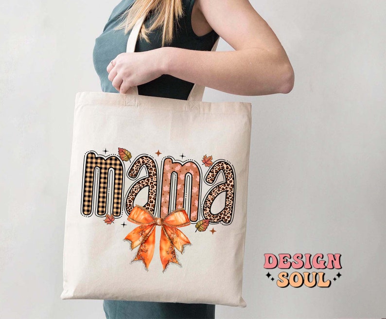 May include: A white tote bag with the word "mama" printed in brown, black, and pink patterned letters. The letters are surrounded by small orange, brown, and black leaves. A large orange bow is centered below the word "mama".