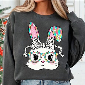 May include: A gray sweatshirt with a colorful cartoon bunny wearing glasses and a bow. The bunny has pink ears with white polka dots, a white bow with black polka dots, and black-rimmed glasses with colorful floral accents.